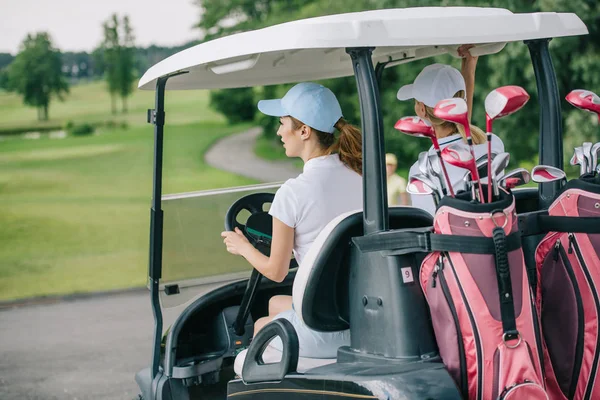 Rückansicht von Golferinnen in Mützen, die Golfkarren mit Ausrüstung auf dem Golfplatz fahren — Stockfoto