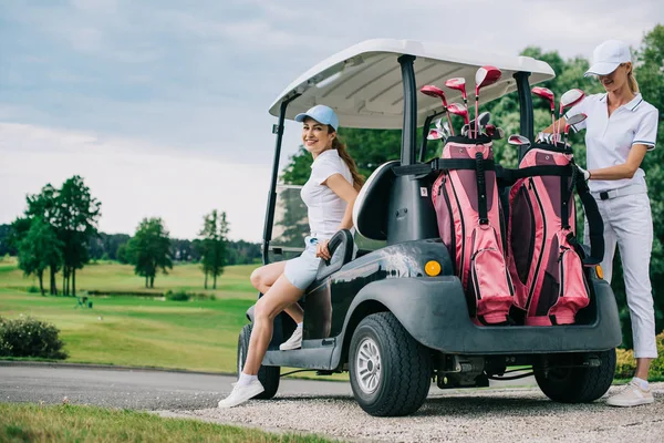 Улыбающиеся женщины-гольфистки на гольф-каре готовятся к игре на поле для гольфа — стоковое фото