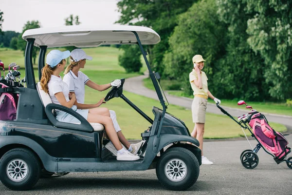 Vista lateral de golfistas femeninas en gorras en carrito de golf y amigo hablando en smartphone en campo de golf - foto de stock