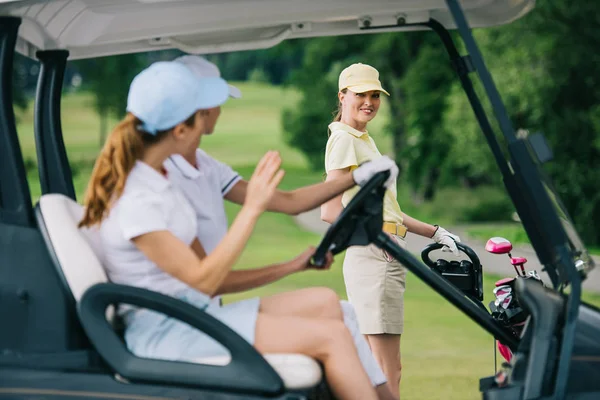 Foco seletivo de golfistas do sexo feminino em bonés no carrinho de golfe cumprimentando amigo com equipamento de golfe no campo de golfe — Fotografia de Stock