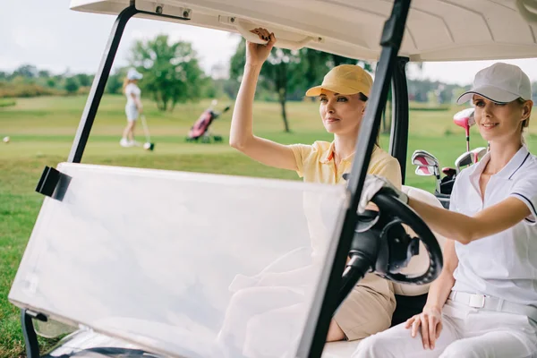 Smiling female golfers in caps riding golf cart at golf course — Stock Photo
