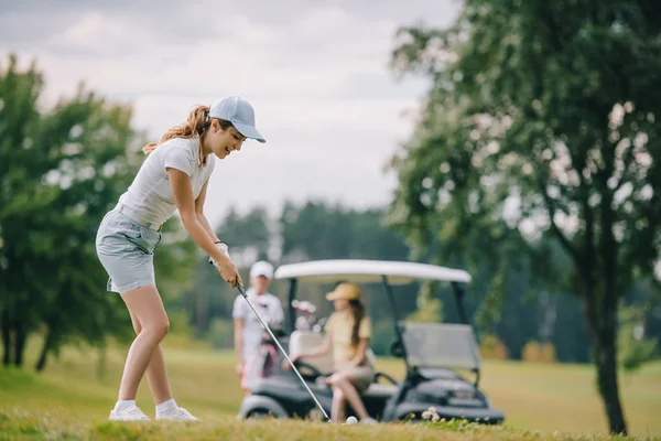 Foyer sélectif de la femme avec le club de golf jouant au golf et des amis se reposant à la voiturette de golf sur la pelouse verte — Photo de stock