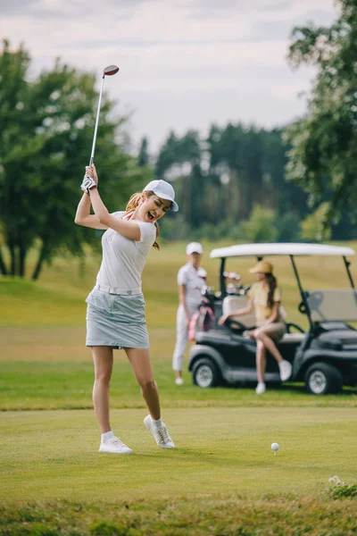 Foco seletivo da mulher com clube de golfe jogando golfe e amigos descansando no carrinho de golfe no gramado verde — Fotografia de Stock