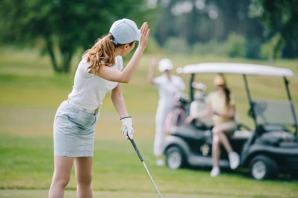 Frau mit Golfschläger winkt Freunden am Golfcart auf grünem Rasen zu — Stockfoto