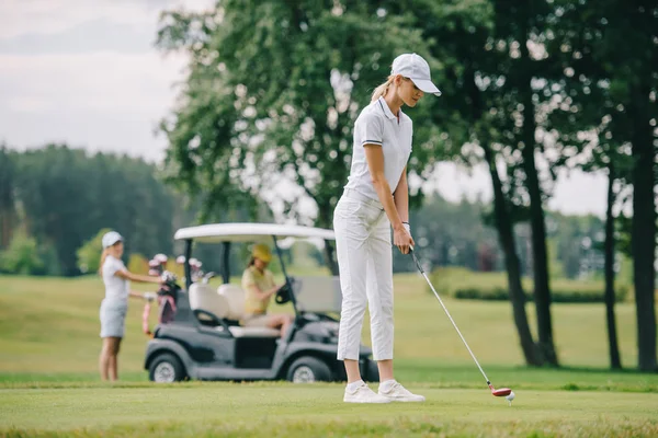 Selective focus of woman with golf club playing golf and friends resting at golf cart on green lawn — Stock Photo