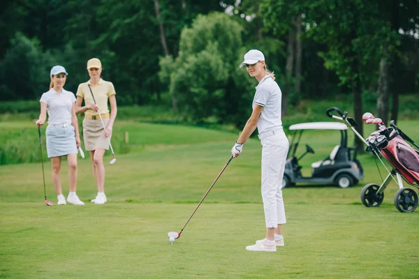 Selektiver Fokus der Frau in Mütze beim Golfspielen, während Freunde in der Nähe auf dem Golfplatz stehen — Stockfoto