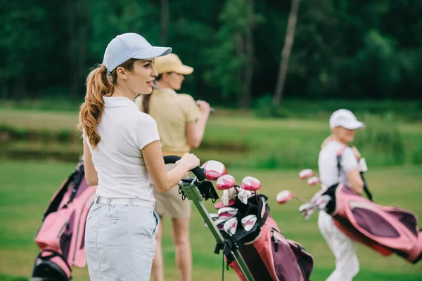 Селективное внимание женщин в шапках с снаряжением для гольфа, идущих по зеленой лужайке на поле для гольфа — стоковое фото