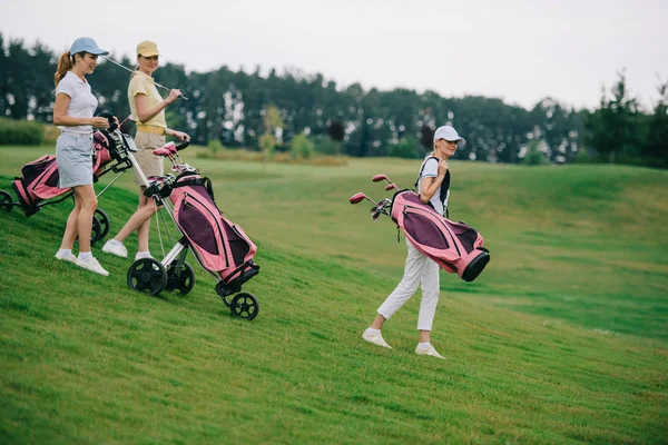 Вид сбоку на женщин в полосах и кепках с экипировкой для гольфа, прогуливающихся по зеленой лавке на поле для гольфа — стоковое фото