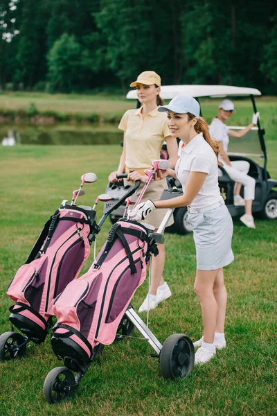 Foco seletivo de jogadores de golfe do sexo feminino com equipamento de golfe e amigo no carrinho de golfe atrás no gramado verde — Fotografia de Stock