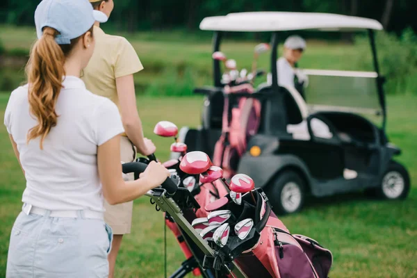 Enfoque selectivo de las jugadoras de golf con equipo de golf y amigo en carrito de golf detrás en césped verde - foto de stock