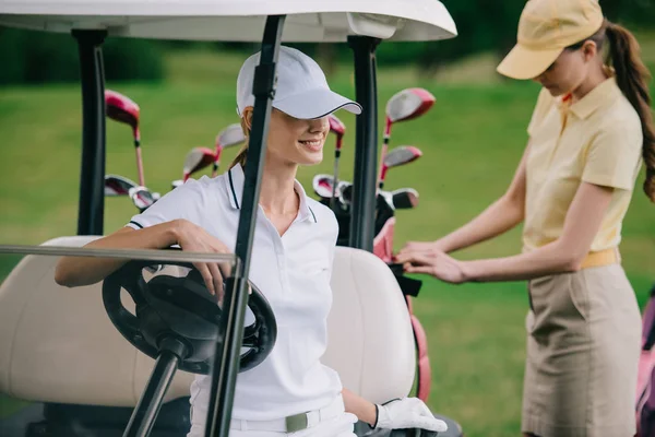 Foco seletivo de jogadores de golfe do sexo feminino em bonés no carrinho de golfe no campo de golfe — Fotografia de Stock