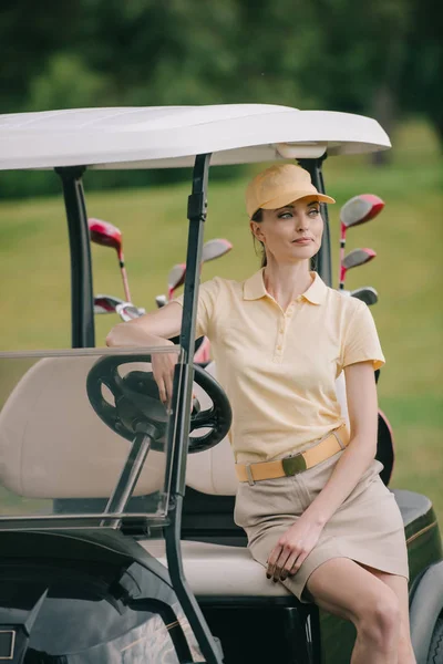 Portrait de femme souriante en polo et casquette assise sur une voiturette de golf et regardant ailleurs — Photo de stock