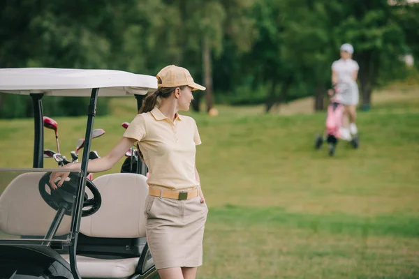 Selektiver Fokus der Golfspielerin in Cap am Golfcart auf grünem Rasen — Stockfoto