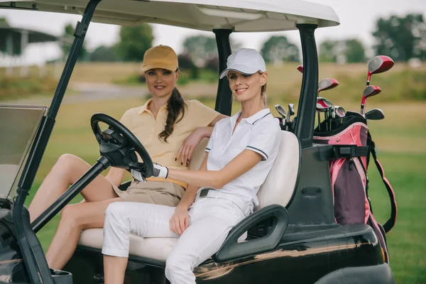 Mujeres sonrientes en gorras sentadas en el carrito de golf y mirando a la cámara - foto de stock