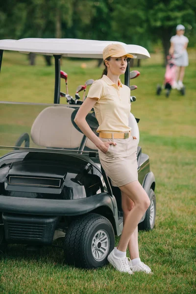 Selective focus of female golf player with hands in pockets at golf cart on green lawn — Stock Photo