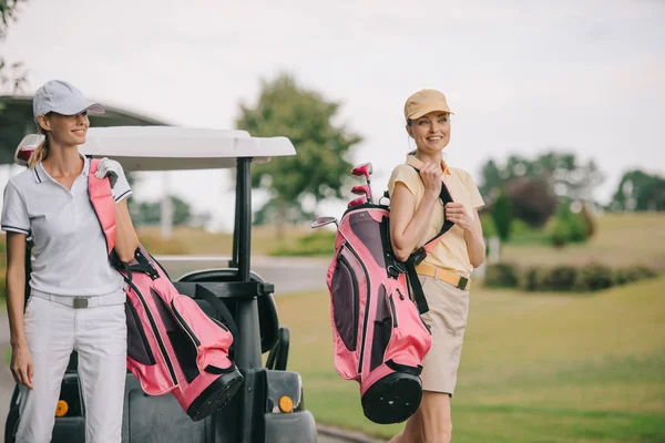 Mujeres sonrientes en polos y gorras con equipo de golf en el campo de golf en el día de verano - foto de stock