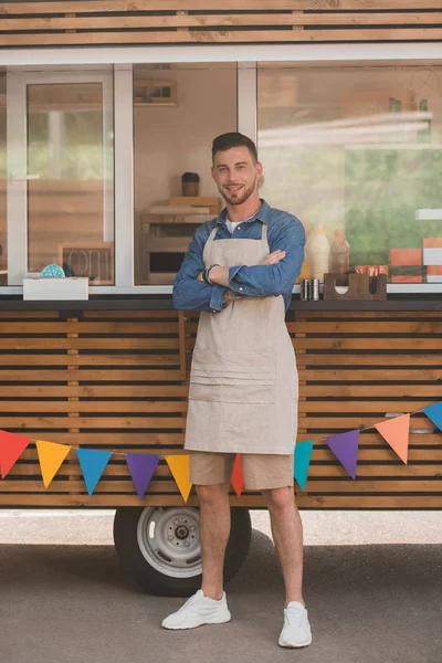 Beau jeune homme dans le tablier souriant à la caméra tout en se tenant avec les bras croisés près du camion de nourriture — Photo de stock