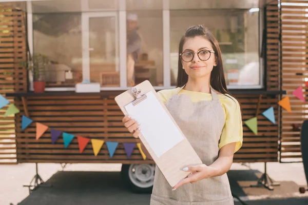 Красивая молодая женщина в фартуке и очках держит пустой планшет и улыбается в камеру, стоя рядом с фургоном с едой — стоковое фото
