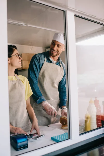 Joven hombre y mujer sonriendo entre sí mientras trabajan juntos en camión de comida - foto de stock