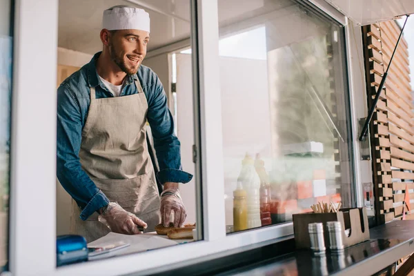 Beau jeune chef souriant regardant loin tout en cuisinant dans un camion de nourriture — Photo de stock