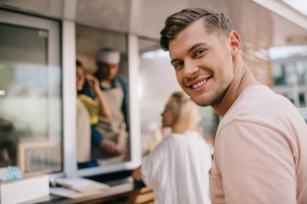 Joven guapo sonriendo a la cámara mientras está de pie en la cola en camión de comida - foto de stock