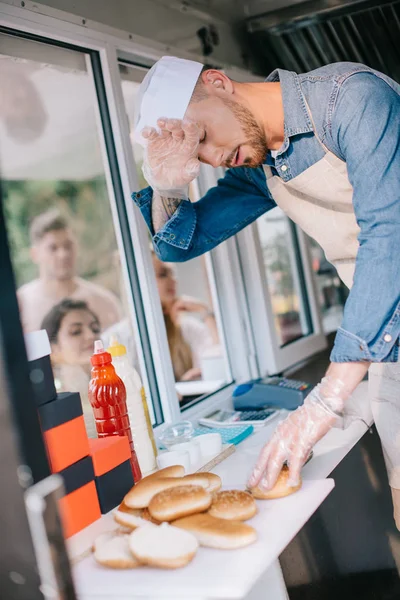 Chef cansado trabajando en camión de comida mientras los clientes esperan fuera - foto de stock