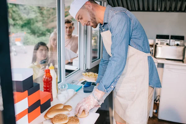 Jeune chef souriant travaillant dans un camion de nourriture tandis que les jeunes se tiennent à proximité — Photo de stock