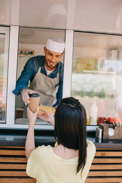 Улыбающийся молодой шеф-повар дает картофель фри и кофе, чтобы пойти к клиентке в фургоне с едой — стоковое фото