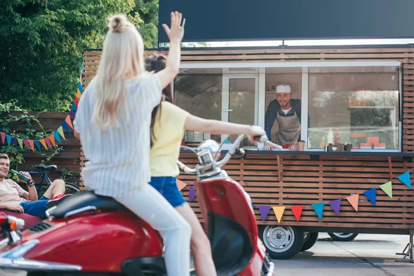 Vista trasera de chica en scooter saludando mano a chef en camión de comida - foto de stock