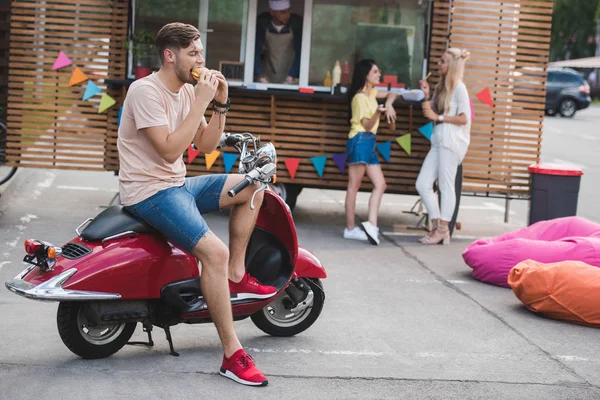 Mann isst Burger und sitzt auf Motorrad in der Nähe von Foodtruck — Stockfoto