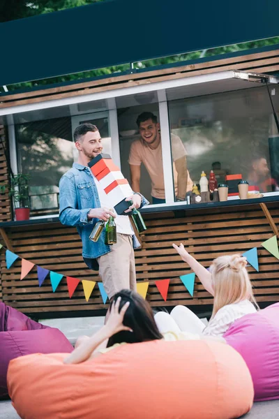 Man carrying boxes with fast food and bottles of beer to friends near food truck — Stock Photo