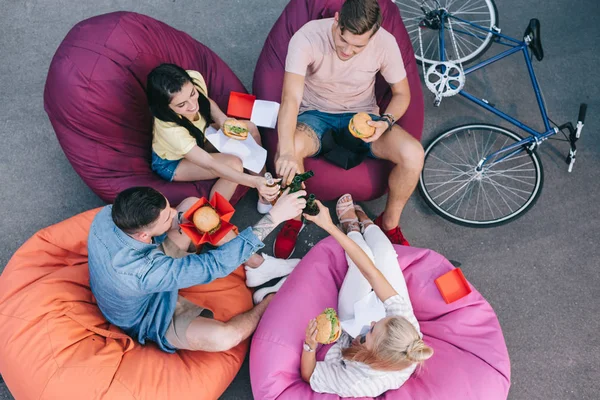 Vue grand angle d'amis cliquetis avec des bouteilles de bière et tenant des hamburgers sur des chaises de sac de haricot — Photo de stock