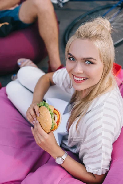 Blickwinkel der attraktiven Frau, die Burger hält und in die Kamera schaut — Stockfoto