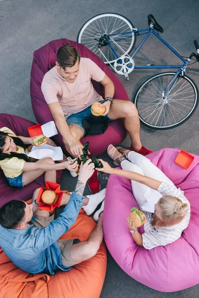 Vue grand angle d'amis cliquetis avec des bouteilles de bière et tenant la restauration rapide sur des chaises de sac de haricot — Photo de stock