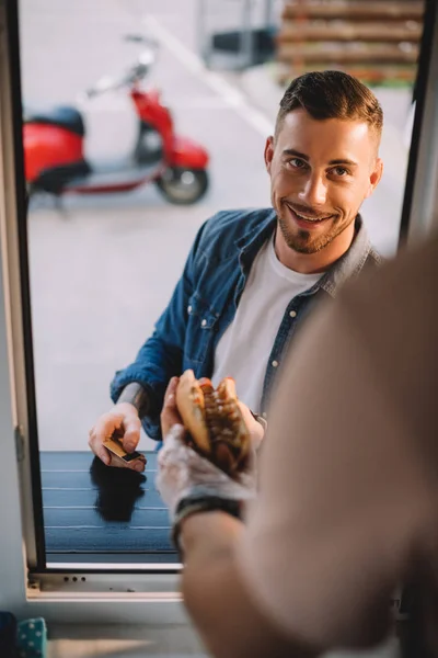 Imagem cortada de chef dando cachorro-quente saboroso ao cliente bonito no caminhão de alimentos — Fotografia de Stock