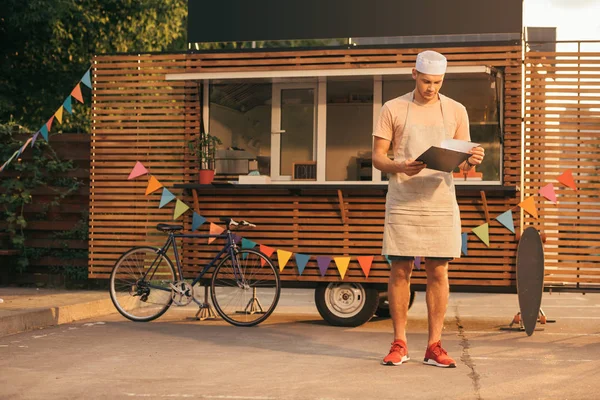 Schöner Koch in Schürze schaut auf Klemmbrett in der Nähe von Food-Truck — Stockfoto