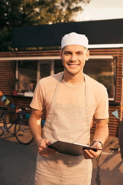 Smiling handsome chef in apron holding clipboard and looking at camera near food truck — Stock Photo