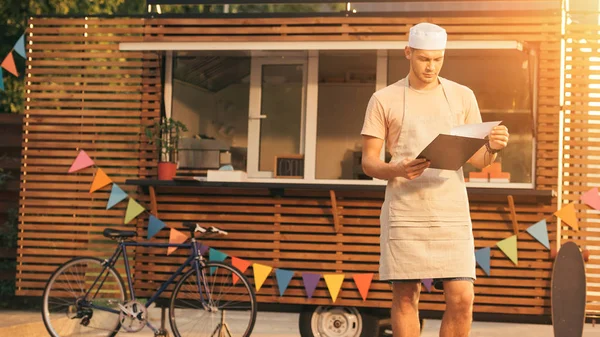 Chef in apron looking at clipboard near food truck — Stock Photo