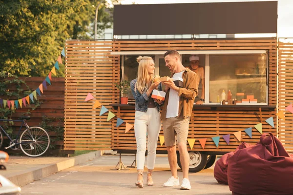 Coppia sorridente che tiene patatine fritte e hamburger vicino al camion cibo — Foto stock