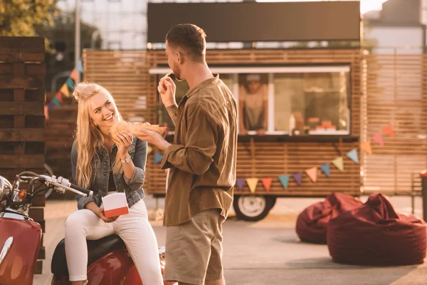 Feliz pareja comiendo papas fritas y hamburguesa cerca de camión de comida - foto de stock