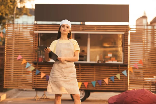 Schöne Köchin in Schürze hält Klemmbrett in der Nähe von Food-Truck und schaut in die Kamera — Stockfoto