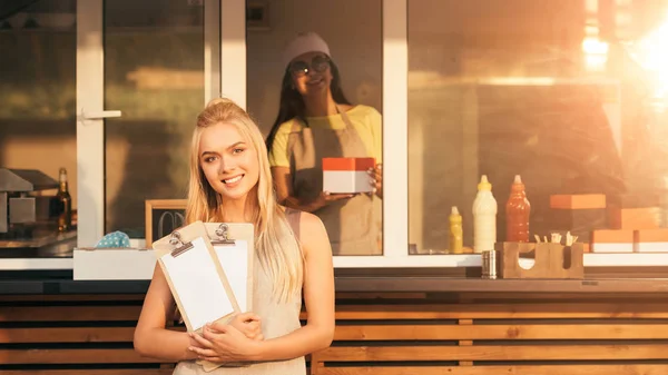 Attraktive lächelnde Köche mit Menü, die in die Kamera neben dem Foodtruck schauen — Stockfoto