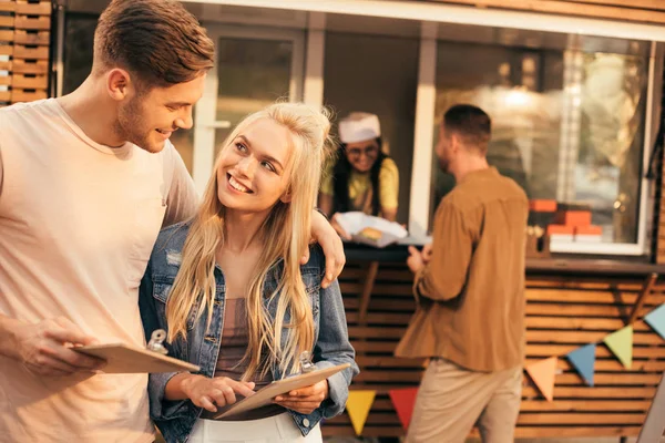 Casal sorrindo segurando menu perto de caminhão de comida — Fotografia de Stock