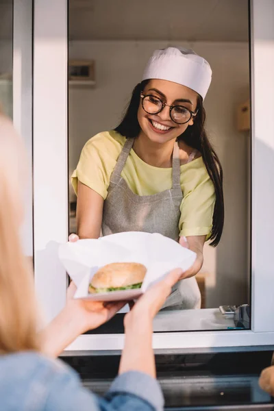 Imagen recortada de chef sonriente dando hamburguesa al cliente en camión de comida - foto de stock