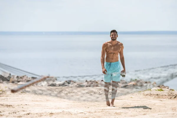 Handsome shirtless man walking on beach, with hammock on foreground — Stock Photo