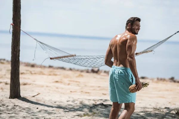 Handsome bearded man holding pineapple and walking on beach with hammock — Stock Photo