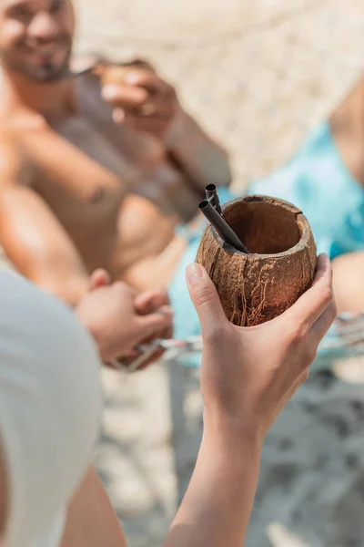 Vista cortada de menina trazendo coquetel de coco para o namorado na rede, foco seletivo — Fotografia de Stock