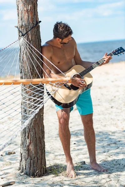 Joven hombre sin camisa tocando la guitarra acústica en la playa cerca de hamaca - foto de stock