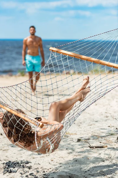 Fidanzata con cocktail di cocco sdraiato su amaca, con fidanzato sullo sfondo, messa a fuoco selettiva — Foto stock