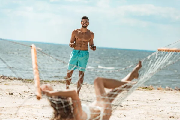 Handsome shirtless man bringing coconut cocktails to his girlfriend on hammock near the sea — Stock Photo